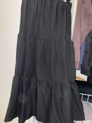 Svart volangkjol med resårmidja - Säljer en svart långkjol med volanger och resår i midjan. Kjolen har en luftig och flowy passform som ger en avslappnad vibe. Perfekt att styla med en crop top eller stickad tröja för en trendig look. Aldrig använt och helt ny!