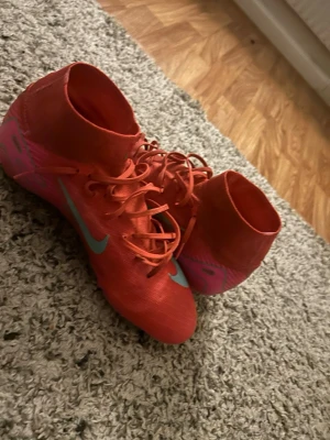 Nike röda fotbollsskor med socka - Säljer ett par röda Nike fotbollsskor med högt skaft och snörning. Skorna har rosa och gröna detaljer samt dobbar för bra grepp på planen. Tillverkade i syntetmaterial och designade för snabbhet och kontroll.