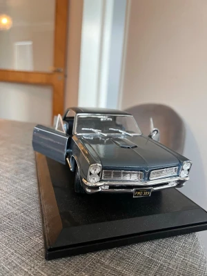 Mörkgrå Pontiac GTO 65 modellbil -  modellbil av en Pontiac GTO i mörkgrå metall med öppningsbara dörrar och motorhuv. Bilen står på en svart displayplatta och har realistiska detaljer som kromade stötfångare, backspeglar och inredning. Perfekt för samlare eller bilentusiaster.