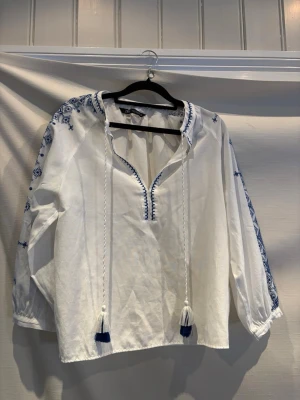 Vit boho-blus med blå broderier Zara - Vit bohemisk blus från Zara med blå broderade detaljer längs ärmar och axlar. Blusen har långa ärmar, snörning med tofsar vid halsen och avslappnad passform. Tillverkad i ett lätt bomullstyg som känns luftigt och skönt.