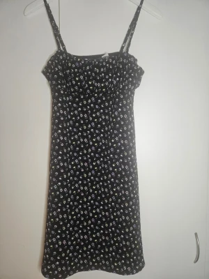 Svart blommig sommarklänning - Söt svart klänning med smala axelband och små blommor i lila, vitt och gult. Klänningen har en smockad överdel och är tillverkad i ett lätt och mjukt material, perfekt för varma dagar. Passformen är normal och längden är kort.