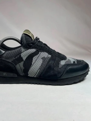 Valentino rockrunners  - Snygga sneakers från Valentino i svart och grått med camo-mönster. Skorna har en mix av mocka, mesh och läderdetaljer samt svarta skosnören. Guldiga detaljer på plösen ger en exklusiv touch. Perfekta för dig som gillar streetwear och unika designer.