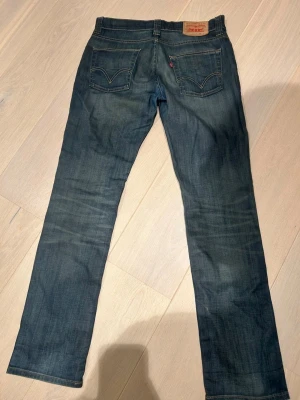 Levi's blå raka jeans - Säljer ett par klassiska blå jeans från Levi's med raka ben och femficksmodell. Jeansen har snygga slitningar och kontrastsömmar samt den ikoniska läderpatchen bak i midjan. Perfekta för en avslappnad och tidlös stil.