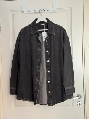 Grå oversized jeansskjorta cubus - Snygg grå oversized jeansskjorta från cubud med klassisk krage och knappar framtill. Skjortan har långa ärmar och är tillverkad i ett robust denimtyg som ger en cool och avslappnad look. Perfekt att bära öppen eller stängd.