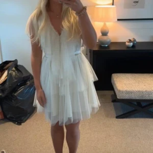 Vit kortklänning från Ida Sjöstedt - Superfin vit kortklänning från Ida Sjöstedt med lager på lager av tunn tyll, v-ringning och volangdetaljer. Klänningen har korta ärmar och ett knytband i midjan som ger en romantisk och luftig känsla. Perfekt för dig som vill sticka ut med en drömmig look. Aldrig använd