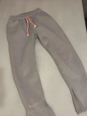 Syna world - Säljer ett par grå mjukisbyxor från SikSilk med rosa dragsko i midjan och stort rosa graffiti-tryck på baksidan. Byxorna har broderad logga vid benet och är tillverkade i mjukt, bekvämt material. Perfekta för en chill och avslappnad stil.