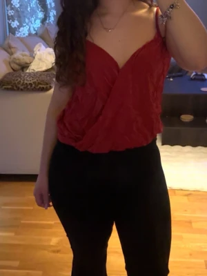 Röd omlott linnetopp med smala band - Snygg röd omlott linnetopp med smala axelband och djup v-ringning. Toppen har ett lätt glansigt tyg och en avslappnad passform som ger en chic vibe. Perfekt att styla med höga jeans eller kjol för en trendig look.