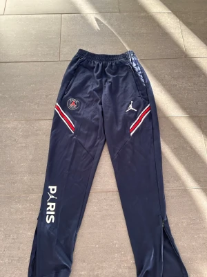 PSG x Jordan mörkblå träningsbyxor - Mörkblå träningsbyxor från PSG x Jordan med vita och röda detaljer vid fickorna, PSG-logga och Jumpman-logga framtill samt PARIS-tryck på ena benet. Byxorna har resår i midjan och dragkedjor vid bensluten. Perfekta för träning eller chill. Storlek S men passar även M skick 8/10
