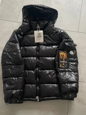 Svart Moncler dunjacka med huva - Snygg svart dunjacka från Moncler med glansig finish och klassisk logga på ärmen. Jackan har huva, dragkedja framtill och tryckknappar vid ärmslut. Perfekt för kalla dagar och riktigt trendig stil.