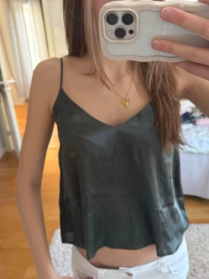 Mörkgrön linnetopp med smala band - Snygg mörkgrön linnetopp med tunna axelband och v-ringad hals. Toppen har en croppad passform och är tillverkad i ett glansigt material som ger en lyxig känsla. Perfekt att styla med jeans eller kjol för en trendig look.