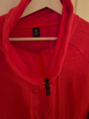 Röd hoodie från SOC med dragkedja - Snygg röd hoodie från SOC i storlek M. Hoodien har dragkedja framtill, justerbar huva med snören och är tillverkad i ett mjukt, sportigt material. Perfekt för dig som gillar en enkel men stilren look.