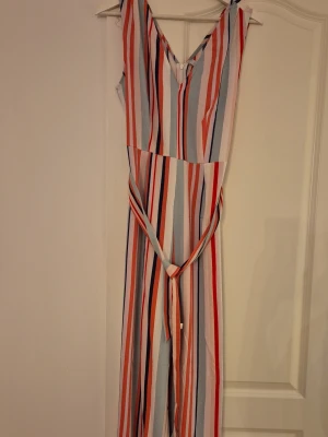 Randig jumpsuit med knytband - Säljer en snygg jumpsuit från NISSA med vertikala ränder i rött, blått, vitt och ljusblått. Modellen har V-ringning, knytband i midjan och dragkedja bak. Perfekt för dig som vill ha en färgglad och trendig look.