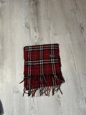 Rutig röd halsduk från burberry - Snygg röd halsduk från Burberry med klassiskt rutmönster i svart, vitt och beige. Halsduken har fransar i ändarna och är tillverkad i mjukt material som känns skönt mot huden. Perfekt accessoar för att lyfta din outfit.