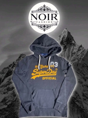 Mörkblå Superdry hoodie med tryck - Mörkblå hoodie från Superdry med stor gul och vit logga på bröstet. Klassisk känguruficka framtill, vita dragsnören i huvan och ribbade muddar. Perfekt för en avslappnad streetwear-look.