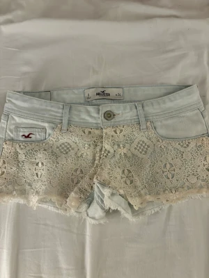 Ljusblå spetsdetaljerade shorts Hollister - Säljer ett par ljusblå jeansshorts från Hollister med snygga spetsdetaljer framtill. Shortsen har klassisk femficksdesign, knappgylf och bälteshällor. Perfekta för varma dagar och ger en romantisk touch till din outfit.