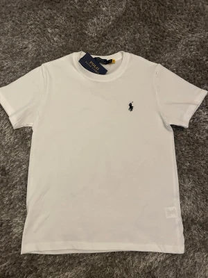 Vit t-shirt från Polo Ralph Lauren - Klassisk vit t-shirt från Polo Ralph Lauren med den ikoniska mörkblå loggan broderad på bröstet. T-shirten har rund hals, korta ärmar och är gjord i mjuk bomull för en skön och stilren look.