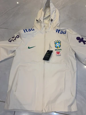 Vit Nike Brasil jacka med huva - Snygg vit träningsjacka från Nike med huva och dragkedja. Jackan har Brasil-logga, grönt Nike-märke och sponsortryck i blått och lila på bröst och ärmar. Perfekt för dig som gillar fotboll och vill ha en sportig look.