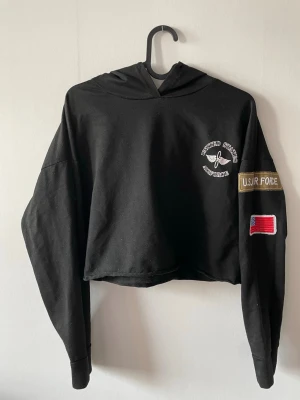 Svart cropped hoodie U.S. Air Force - Svart cropped hoodie med tryck och brodyr. Har huva, vit text och emblem på bröstet samt patchar på ärmen, inklusive amerikansk flagga. Saknar dock snöret till dragskon. 