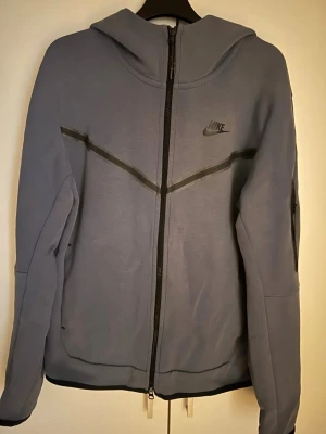 Nike Tech Fleece hoodie  - Nike Tech Fleece hoodie i grått med hel dragkedja och svarta detaljer. Snyggt Nike-logga på bröstet och två sidofickor. Hoodien har en modern passform och är tillverkad i mjukt, bekvämt material. Perfekt för en sportig och avslappnad stil.