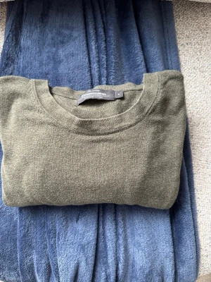 Olivgrön stickad tröja Dressmann - En enkel och stilren olivgrön stickad tröja från Dressmann. Tröjan har rund halsringning, långa ärmar och ribbade muddar vid ärmslut och nederkant. Perfekt att matcha med jeans eller chinos för en avslappnad look.