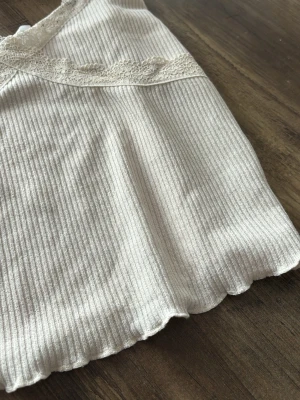 Ljus ribbad linnetopp med spets - Supersöt ljusbeige/krämfärgad ribbad linnetopp med tunna axelband och spetsdetaljer längs v-ringningen och under bysten. Perfekt till sommaren och enkel att styla med jeans eller kjol. Mjuk och stretchig känsla. Den har liten volangdetalj längst ner vilket ger det en söt och fin känsla. Toppen är använd fåtal gånger dock är lappen bortklippt då den var obekväm! 