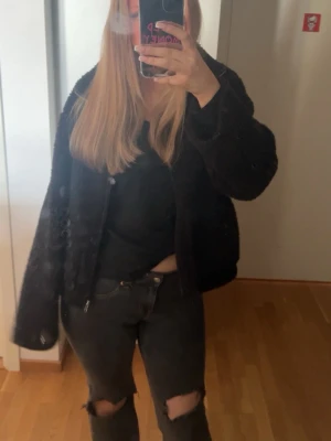 Svart teddyjacka med dragkedja - Mysig svart teddyjacka med dragkedja framtill och avslappnad passform. Jackan har en kortare modell och fluffigt material som ger en trendig look. Perfekt för att hålla värmen och samtidigt se snygg ut. Passar till jeans och sneakers för en cool vardagsstil.