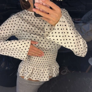 Vit prickig blus med smock - Supersöt vit blus med mörkbruna prickar och smockad midja som ger en snygg siluett. Blusen har långärmade ärmar med lätt utsvängda muddar och rund halsringning. Perfekt för dig som gillar trendiga detaljer och klassiskt mönster.