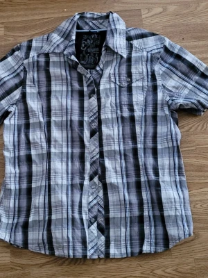 Rutig kortärmad skjorta grå/svart - Kortärmad skjorta från New Sensation i svart, grått och vitt rutigt mönster. Klassisk krage, knappstängning framtill och en bröstficka. Perfekt för dig som gillar en avslappnad men ändå stilren look. Liten i storleken mer en M