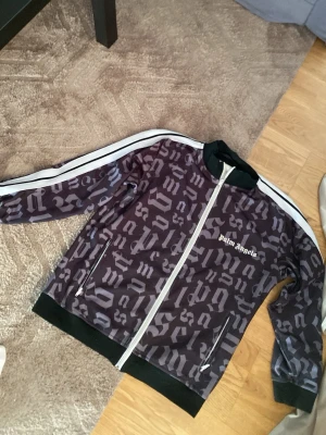 Palm Angels svart zip-tröja - Palm Angels zip-tröja i svart med grått grafiskt mönster och vita detaljer. Tröjan har vita ränder längs ärmarna, ribbade muddar och ståkrage. Märkets logga är tryckt på bröstet. Perfekt för en streetwear-look.