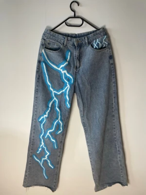 Blå jeans med blixtmotiv - Unika blå jeans med bred passform och handmålat blixtmotiv i ljusblått och vitt på vänster ben och ficka. Klassisk femficksmodell i denim med råa kanter vid benslut. 