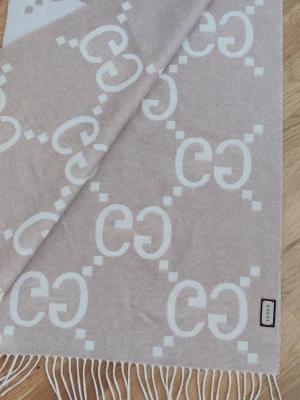Beige halsduk med GG-mönster  - Snygg beige halsduk med ikoniskt GG-mönster i vitt och dekorativa fransar i kanterna. Ull.