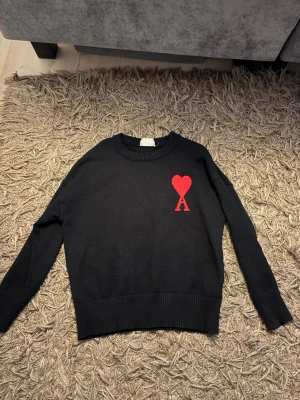 Svart stickad tröja Ami Paris - Svart stickad tröja från Ami Paris med röd hjärtlogga och bokstaven A broderad på bröstet. Tröjan har rund halsringning, långa ärmar och ribbade muddar vid ärmslut och nederkant. Perfekt för en stilren och trendig look. Nypris ca 3500kr