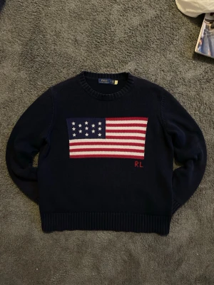 Stickad Ralph Lauren tröja - Mörkblå stickad tröja från Polo Ralph Lauren med amerikansk flagga i rött, vitt och blått på bröstet samt RL-broderi. Tröjan har rund halsringning och ribbstickade muddar vid ärmslut och nederkant. Perfekt för dig som gillar klassisk preppy stil.