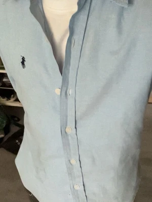 Ljusblå skjorta från Ralph Lauren - Klassisk ljusblå skjorta från Ralph Lauren med broderad logga på bröstet. Skjortan har lång ärm, knappar framtill och en tidlös krage. Tillverkad i mjuk bomull för en bekväm känsla och snygg passform.