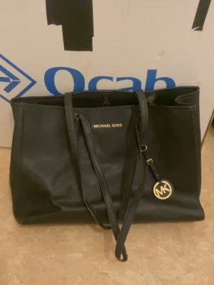 Svart Michael Kors handväska - Snygg svart handväska från Michael Kors i rektangulär modell med guldfärgade detaljer och logotyp. Väskan har dubbla handtag, dragkedja och flera fack inuti för smart förvaring. Tillverkad i slitstarkt konstläder och har en klassisk, stilren look. Vid snabb affär kan priset diskuteras 