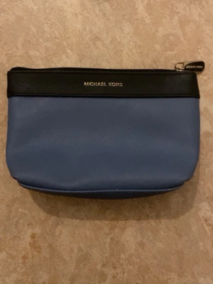 Blå nessesär från Michael Kors - Snygg clutch från Michael Kors i blått konstläder med svart kant upptill och guldfärgad logga. Väskan har dragkedja med metall-detalj och en stilren, enkel design som passar till många olika looks.