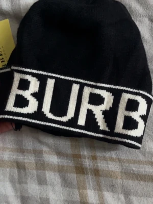 Svart Burberry mössa med logga - Säljer en svart mössa från Burberry med bred uppvikt kant och stor vit logotyp runt hela mössan. Klassisk och stilren design i mjukt stickat material som håller dig varm. Perfekt accessoar för dig som gillar exklusiva märken och streetstyle.