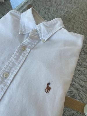 Polo Ralph Lauren Skjorta Vit - Klassikt vit skjorta från Polo Ralph Lauren  Skick: Fint, liten skada baktill, Se bild 4 - Plagget är prissatt därefter.   Mått: • Axel - Axel: 44cm • Axel - Midja: 60cm • Armhåla - Armhåla: 46cm • Axel - Ärmslut: 61cm  Detaljer • Tillverkad i 100% Bomull • -Liten skada baktill, Se bild 4 • Broderad logga på vänster sida av bröstet • Luktfri  Nypris: 1995kr - Mitt Pris: 349kr Skickas inom 24 timmar efter köp!