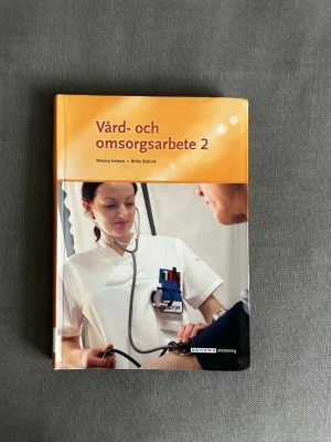 Vård- och omsorgsarbete 2 bok - Kurslitteratur med titeln 'Vård- och omsorgsarbete 2' av Monica Imborn och Britta Åsbrink. Boken har ett orange omslag med bild på vårdpersonal och används inom vård- och omsorgsutbildningar.