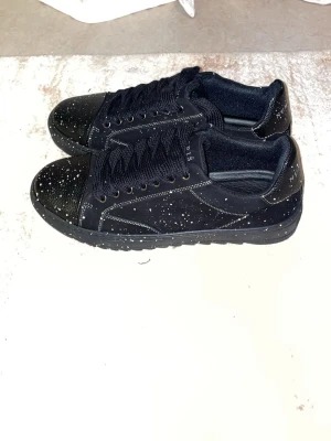 Svarta sneakers med vita stänk - Snygga svarta sneakers från Sandro Paris med vita stänk över hela skon. Skorna har låga skaft, svarta skosnören och en klassisk siluett. Materialet är mocka och textil, vilket ger en cool och modern look.