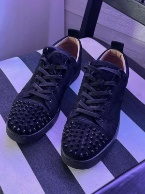 Svarta sneakers med nitar - Säljer ett par svarta sneakers från Christian Louboutin med mattsvarta nitar på tån och svart mocka över hela skon. Skorna har svarta skosnören, låg profil och en klassisk gummisula. Insidan är beige och de har en cool, edgy vibe.