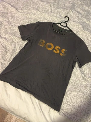 Grå BOSS t-shirt med gul text - Snygg grå t-shirt från BOSS i storlek XS. T-shirten har korta ärmar och ett stort gult BOSS-tryck på bröstet. Tillverkad i mjuk bomull som känns skön mot huden. Perfekt för dig som gillar stilrena och klassiska plagg med en twist. XS men stor i storlek så passar S