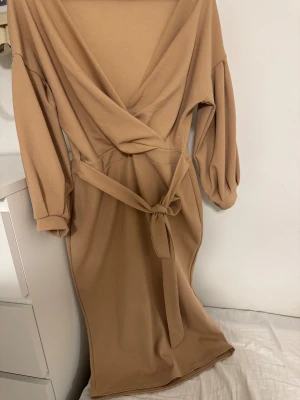 Beige omlottklänning med bälte - Stilren beige omlottklänning från SHEIN med v-ringning och knytbälte i midjan. Klänningen har långa ärmar och en mjuk, lätt struktur i tyget. Perfekt för dig som gillar enkel men snygg design.