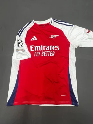 Arsenal matchtröja Adidas röd/vit - Snygg Arsenal matchtröja från Adidas i rött och vitt med blå detaljer. Tröjan har klubbmärke, Adidas-logga och Emirates Fly Better-tryck på bröstet samt Champions League-märke på ärmen. Tillverkad i lätt och ventilerande material, perfekt för fotboll.