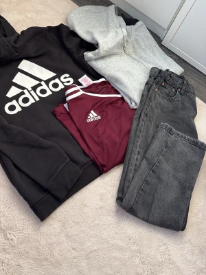 Adidas hoodie och t-shirt, jeans - Säljer en svart hoodie med stor vit Adidas-logga, en vinröd Adidas t-shirt med vita detaljer, ett par grå jeans med fem fickor och en ljusgrå zip-hoodie. Perfekt för en avslappnad och sportig stil.