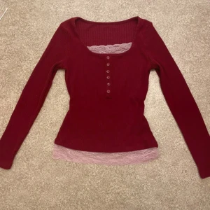Vinröd ribbad topp med spetsdetalj - Vinröd långärmad topp med ribbad struktur och dekorativa knappar framtill. Toppen har en ljusrosa spetskant vid halsringningen och nederkanten som ger en söt touch. Figurnära passform och mjukt material, perfekt för en trendig vardagslook. Hör av er vid frågor!