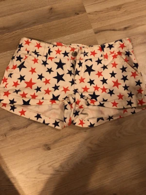 Stjärnmönstrade shorts från Cubus - Säljer ett par vita shorts från Cubus med coolt stjärnmönster i rött och mörkblått. Shortsen har fickor fram och bak, bälteshällor och knappstängning. Perfekta för sommardagar när du vill sticka ut lite extra!
