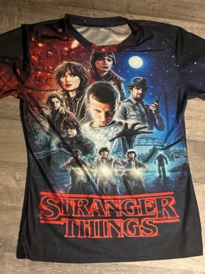 Stranger Things t-shirt med tryck - Cool Stranger Things t-shirt med färgstarkt tryck av karaktärerna och loggan på framsidan. Använd en gång. Trycket går över hela T-shirten fram och bak. 