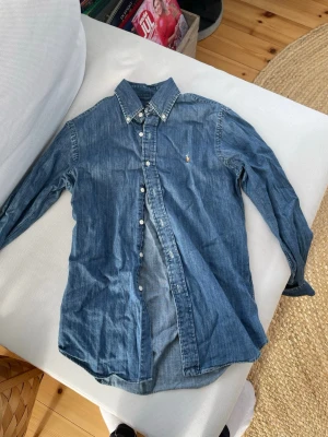 Blå jeansskjorta från Ralph Lauren - Säljer en klassisk blå jeansskjorta från Ralph Lauren med ikonisk broderad logga på bröstet. Skjortan har lång ärm, knappar framtill och en snygg krage. Perfekt till jeans eller chinos för en avslappnad men stilsäker look.
