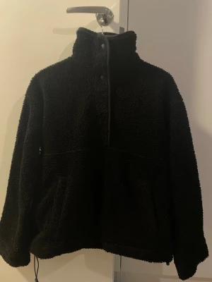 Svart teddyjacka med hög krage - Mysig svart teddyjacka med hög krage och tryckknappar framtill. Jackan har två fickor och dragsko nedtill för justerbar passform. Tillverkad i fluffigt teddy-material som håller dig varm och bekväm. Perfekt för kyliga dagar och enkel att matcha med olika outfits.